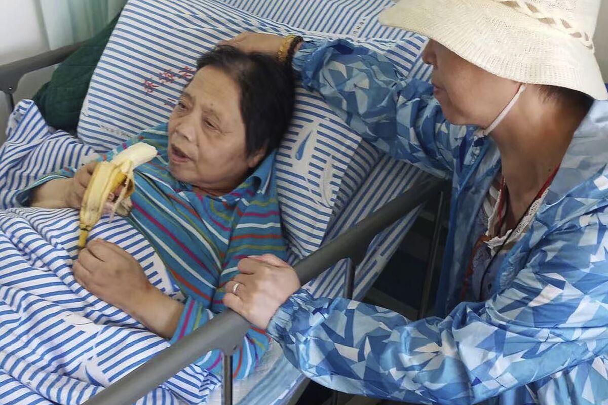 In this photo released by the family of Shen Peiming, Shen Peiming, 71, eats a banana as a family member attends to her at her bed side at the Shanghai Donghai Elderly Care hospital on Sept. 24, 2019. Shen died Sunday morning, April 3, 2022, at the hospital, without her loved ones by her side. Her family, unable to visit because of pandemic restrictions, is unsure of the circumstances of her death. The hospital had reported a COVID-19 outbreak, but Shen had tested negative, as of last week. (Family of Shen Peiming via AP)