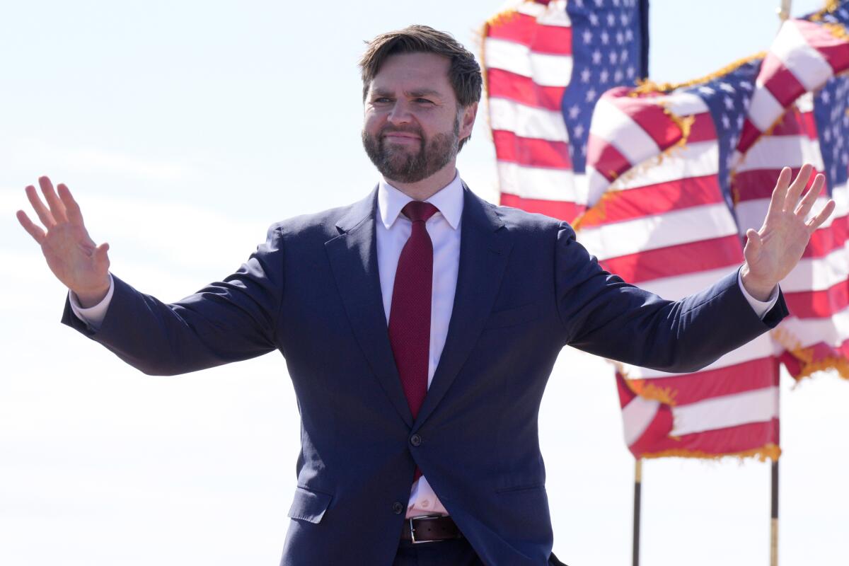 J.D. Vance, wearing a dark suit, stands waving with American flags in background