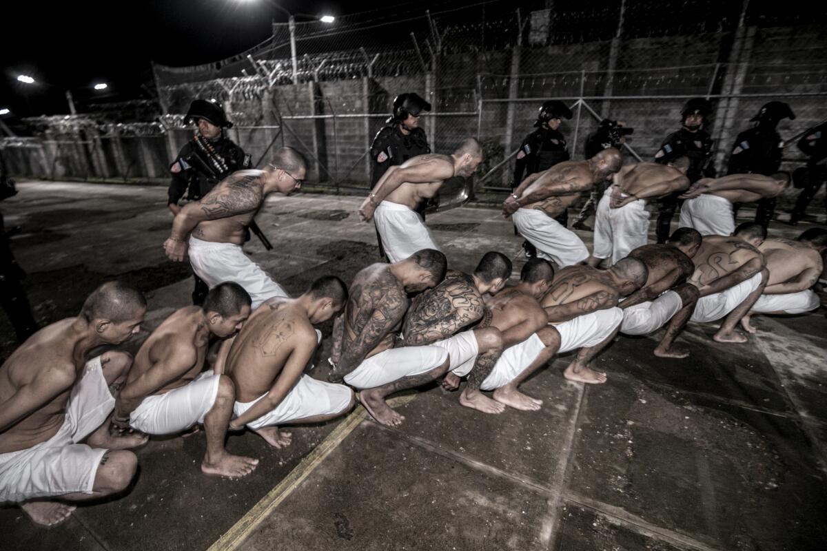 5 men in nothing but shorts run or squat in the dark, hands cuffed behind them, inside a security fence near several officers