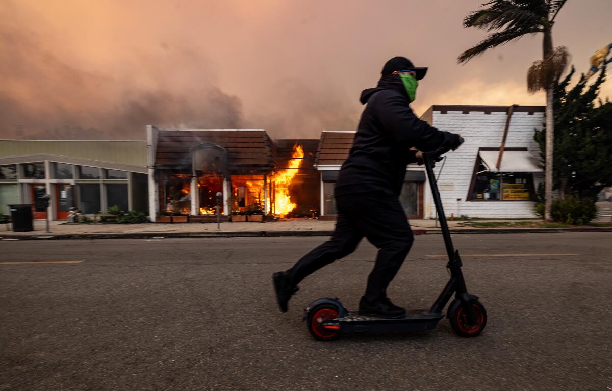A man rides a scooter past a burning business in Palisades Village on Jan. 8.