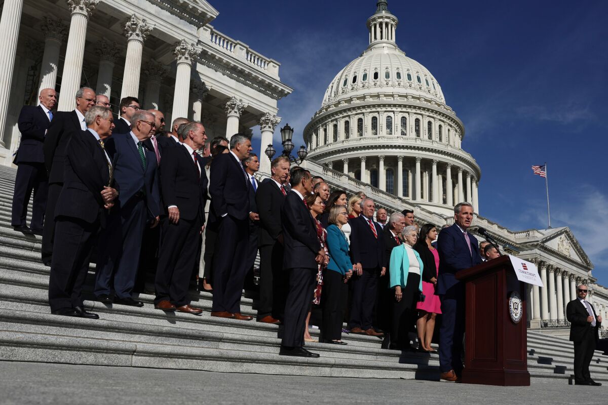A large group standing on a broad stairway behind a man speaking at a lectern, with the U.S. Capitol dome in the background.