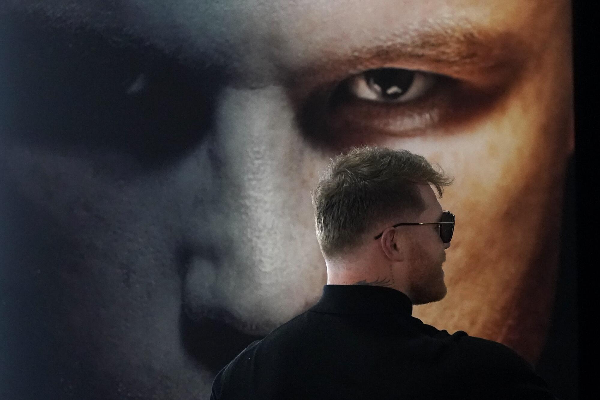 Boxer Canelo Álvarez wears sunglasses and stands facing a poster promoter his fight with Dmitry Bivol.