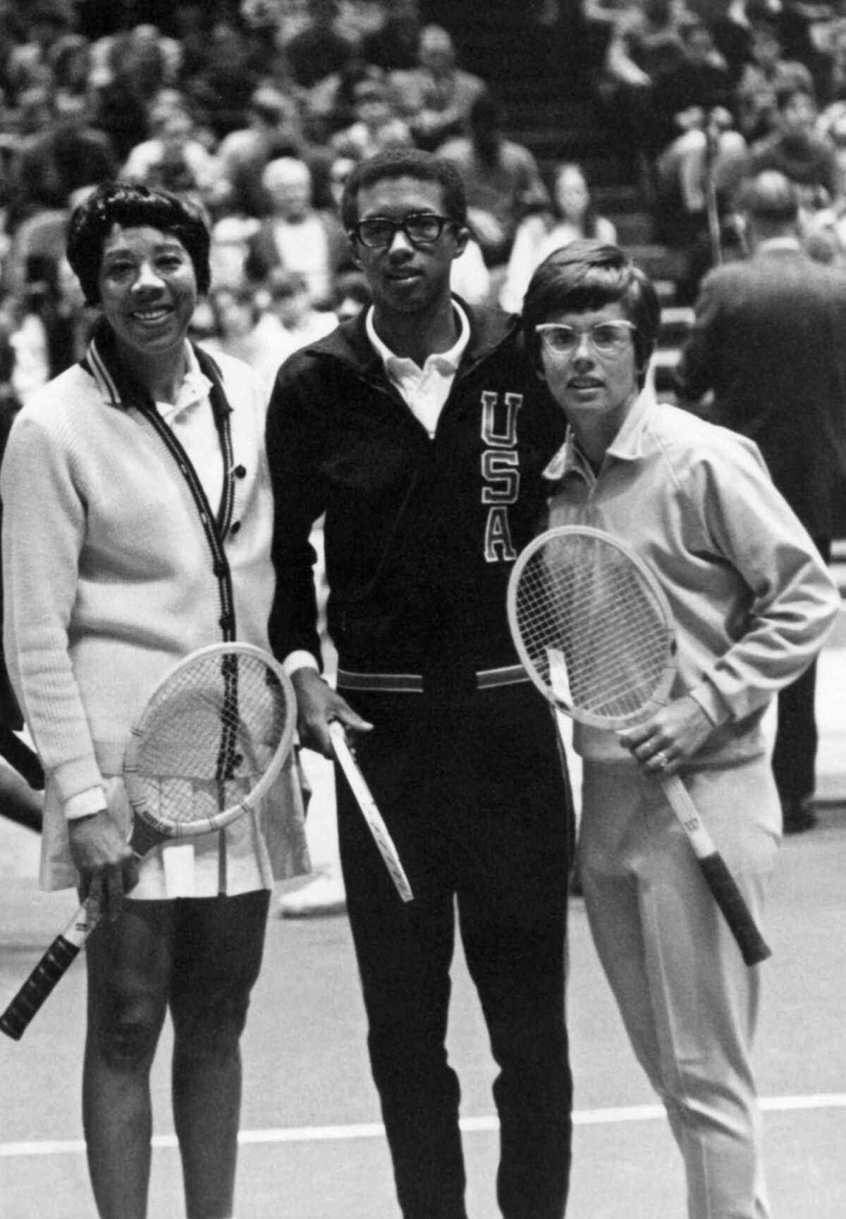 A black-and-white photo of two women with a man in between them, all holding tennis rackets. 