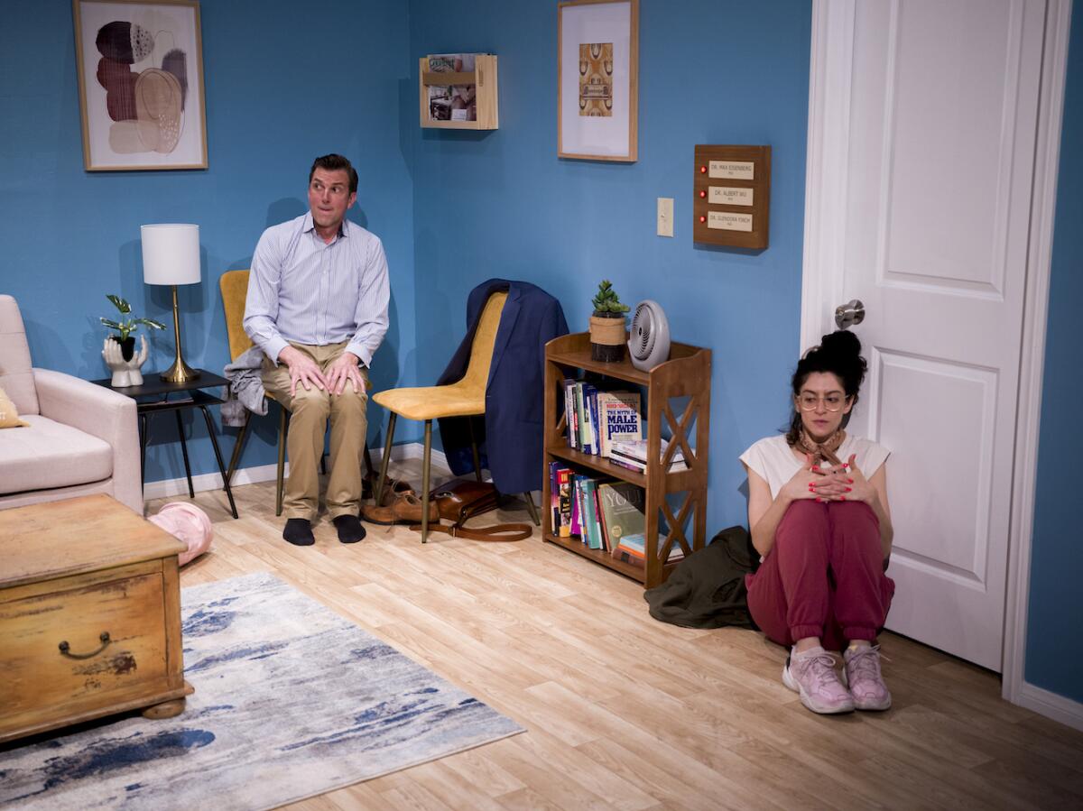 A woman sits on the floor in front of a door while a man sits in a chair in the corner of the room.