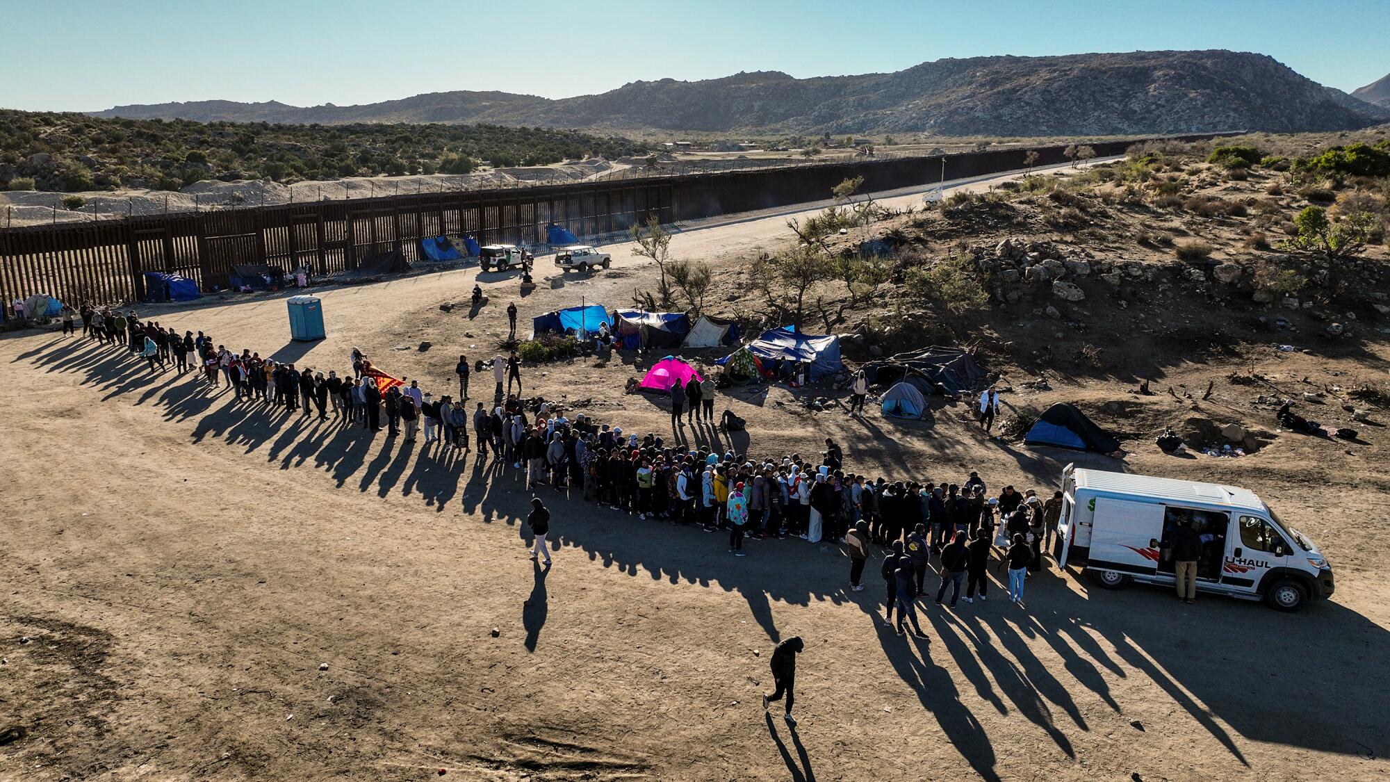 Asylum seekers form a long line behind a vehicle parked in a desert area with a border wall