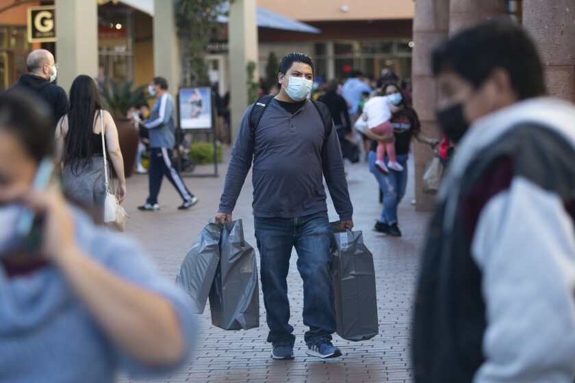 A man wearing a mask carries shopping bags in his hands with other masked people around him
