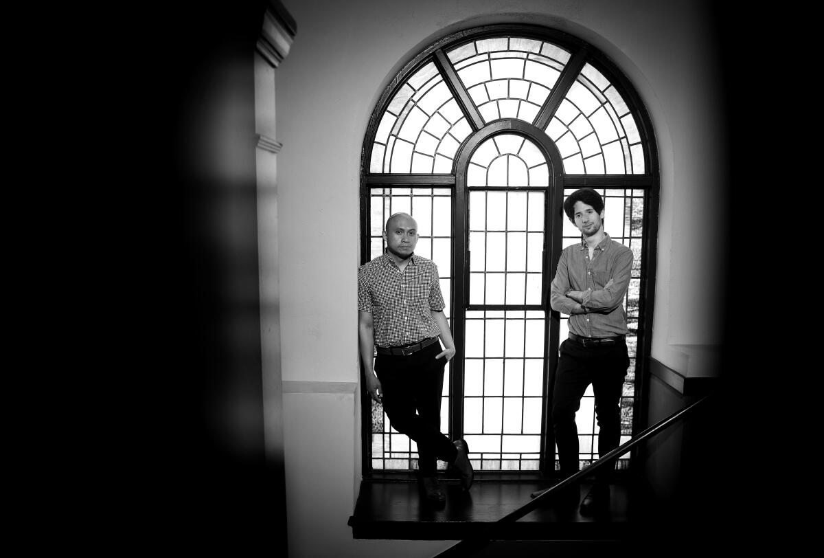 A black and white photograph shows Paulo K Tirol and Noam Shapiro standing before a large arched window.