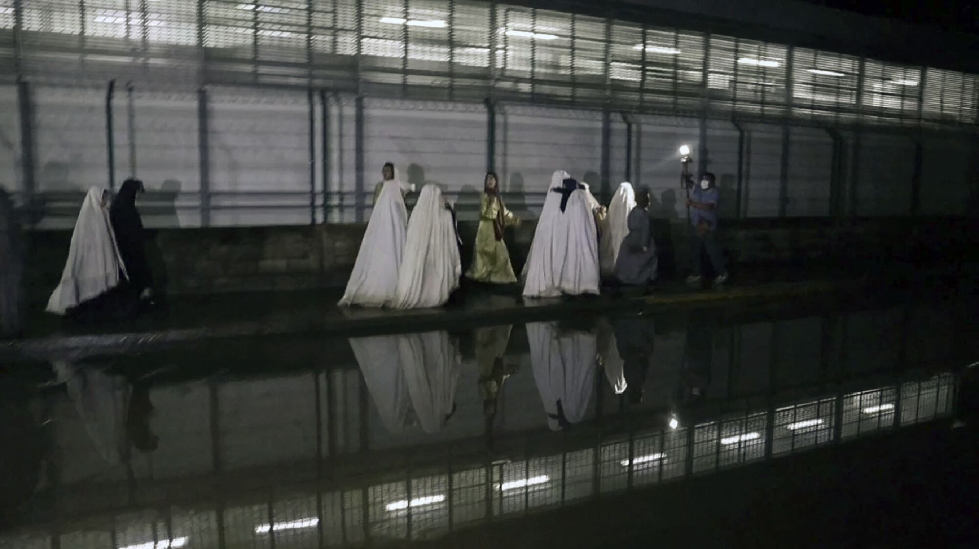 A group people, some cloaked from head to toe in white robes, walk on a sidewalk near a wall with lighting