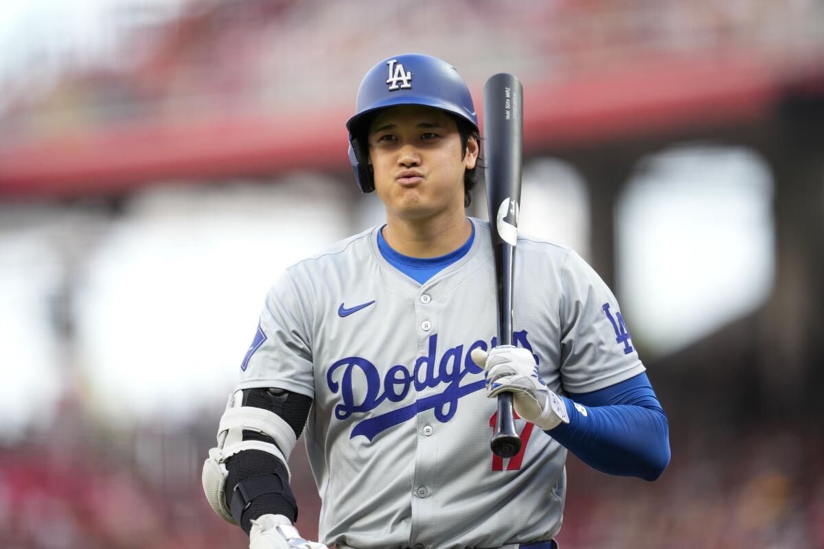Dodgers star Shohei Ohtani, in a batting helmet and gray uniform, returns to the dugout with his bat against his shoulder