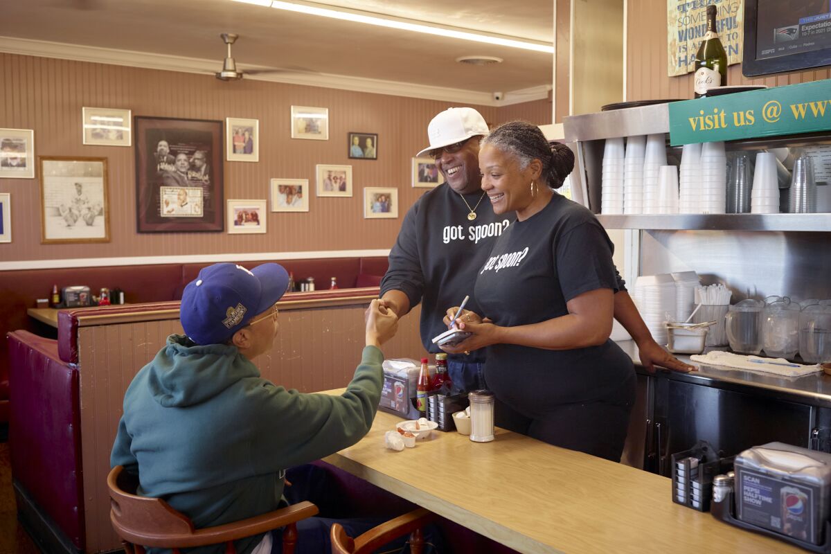 A woman and man stand behind a diner counter, talking with a seated customer