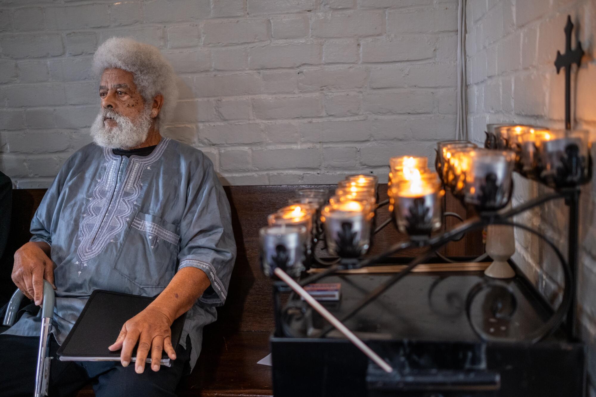 A horizontal frame of a gray-haired man sitting at left and next to a stand with rows of small candles.