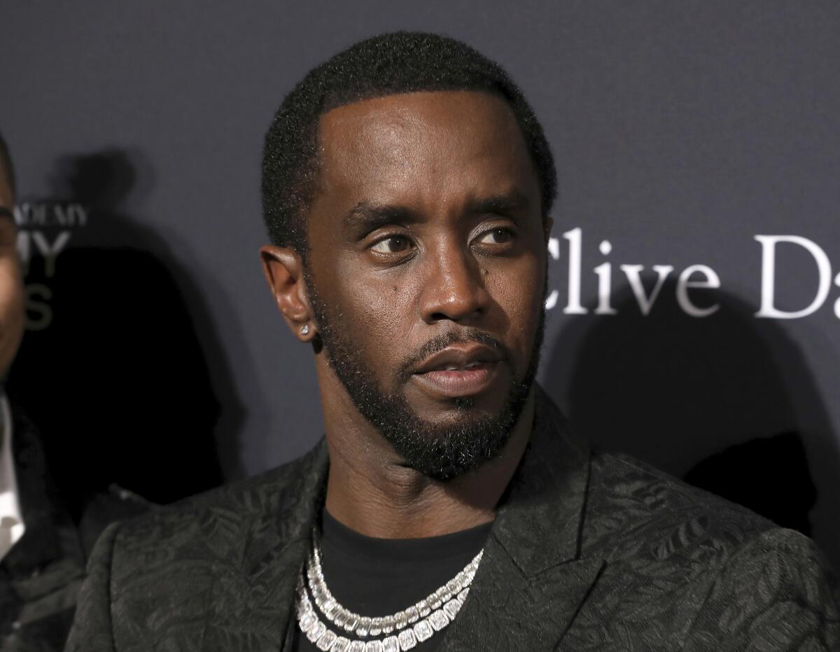 Sean Combs, in a patterned black suit and diamond chains, looks to the side.  