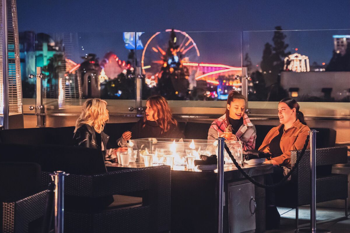 four women at a rooftop bar table with a firepit in the middle and roller coasters lit up  in the background