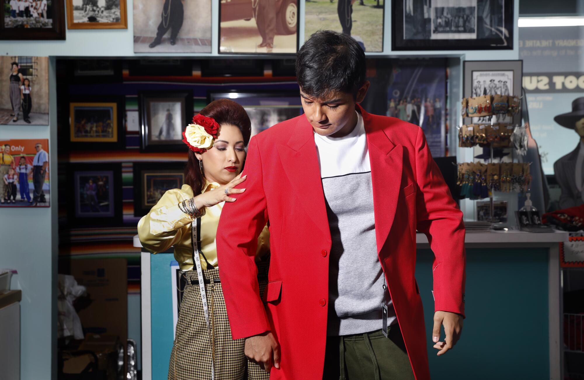 A woman in a 1940s hairdo and yellow shirt fits a teenager in a red zoot suit inside a tailor shop.