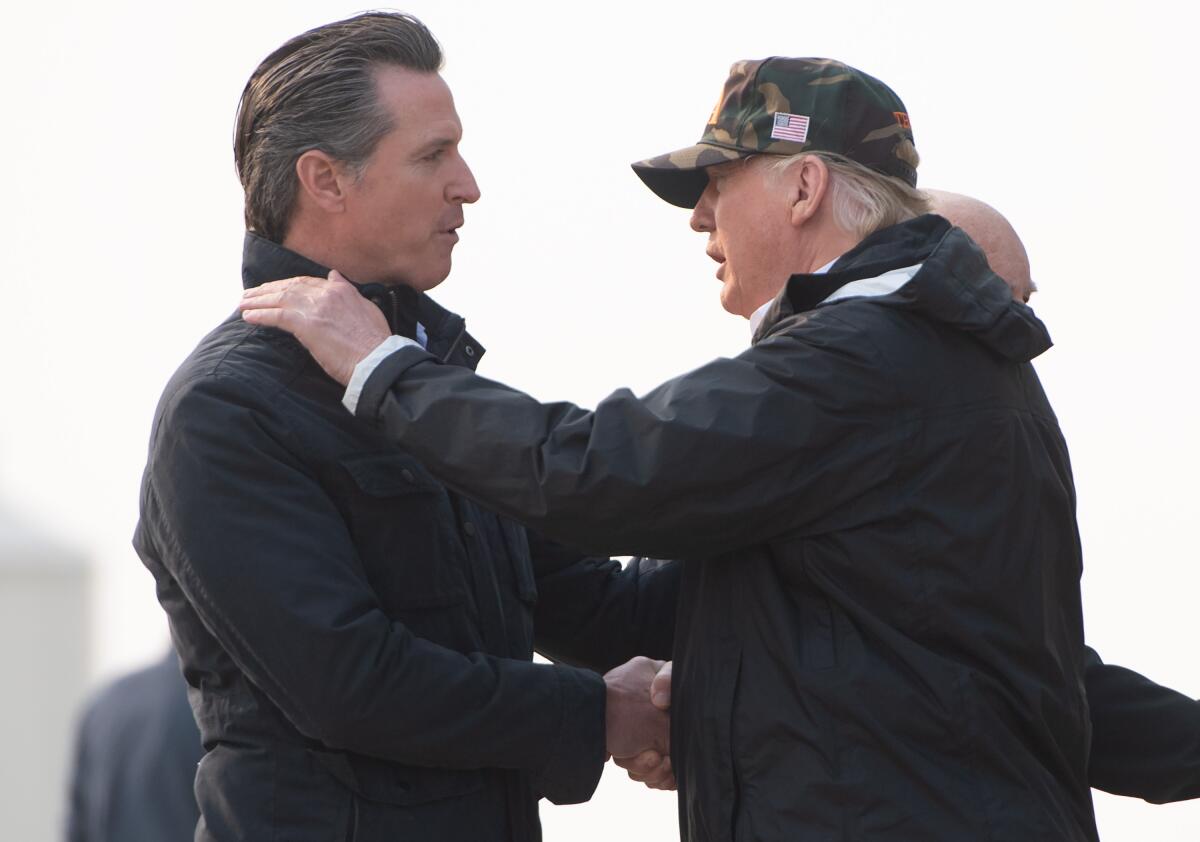 Two men in dark jackets shake hands. The man on the right, in a camouflage cap, has a hand on the other man's shoulder 