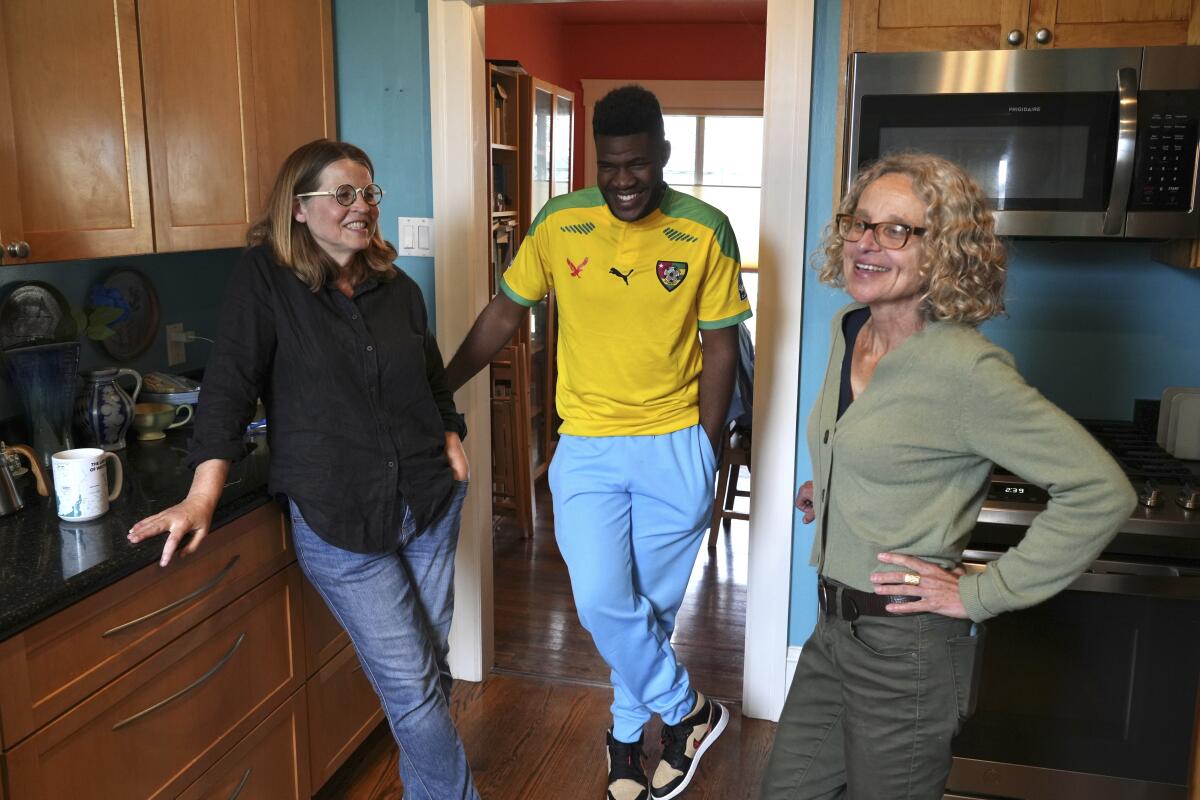 A female couple stand in their kitchen with a smiling man in an athletic jersey.