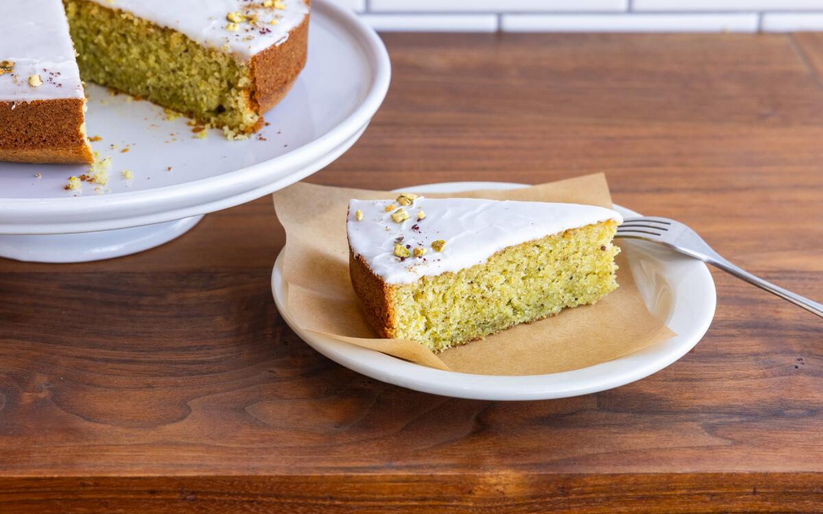 A triangular slice of pistachio olive oil cake with lemon glaze on a small plate next to the remaining cake on a cake stand