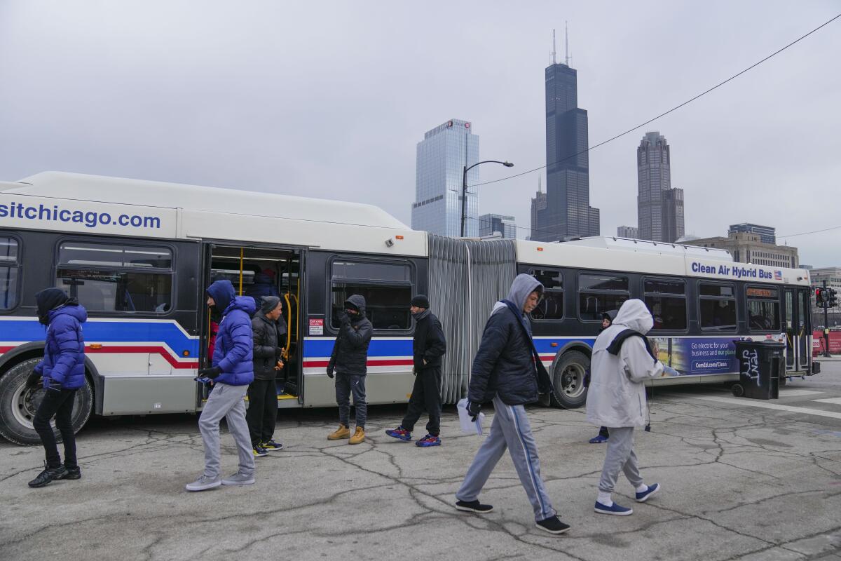 People stand outside buses, with part of a city skyline in the background 