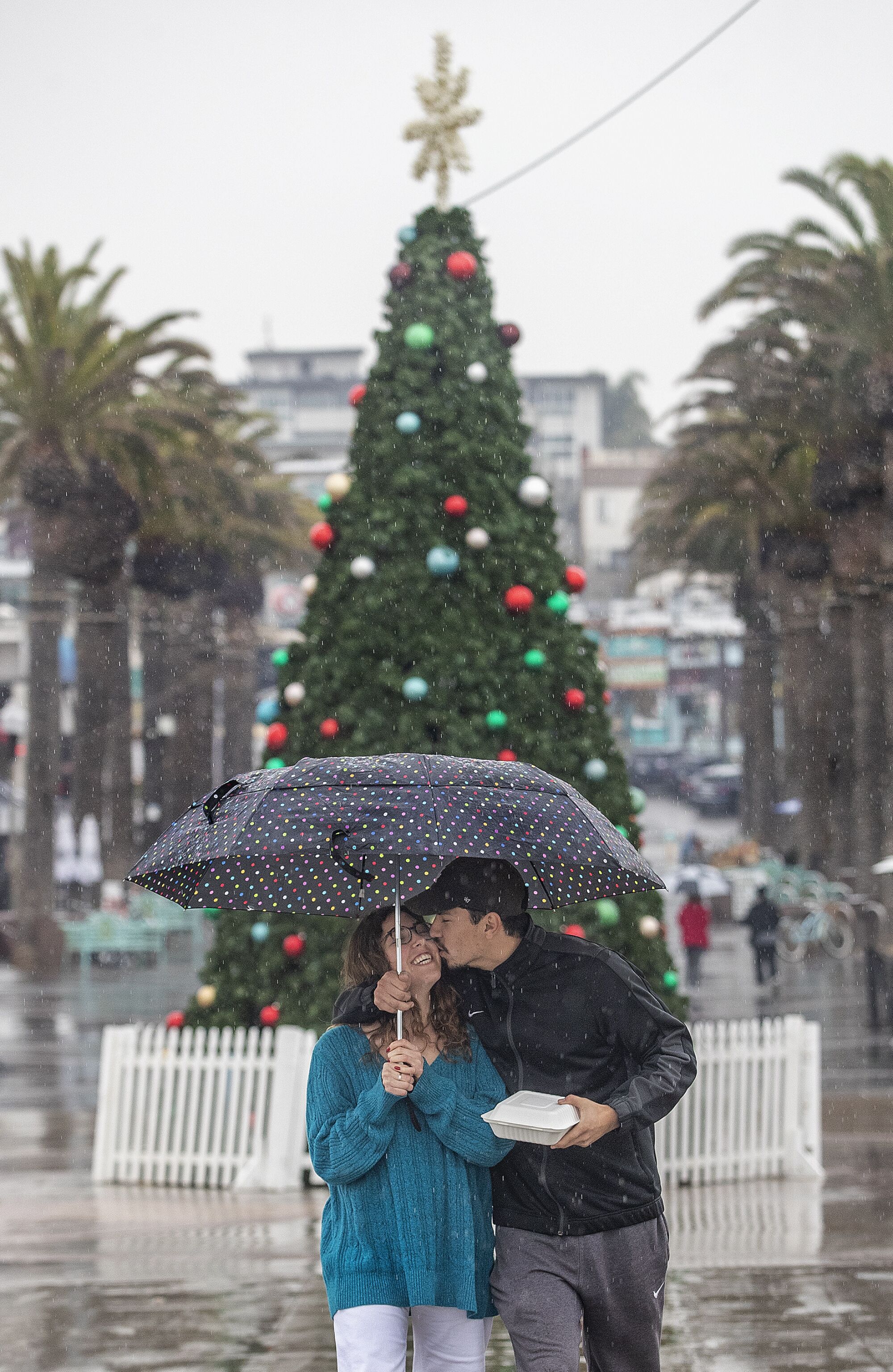A couple share an umbrella in front of an outdoor Christmas tree