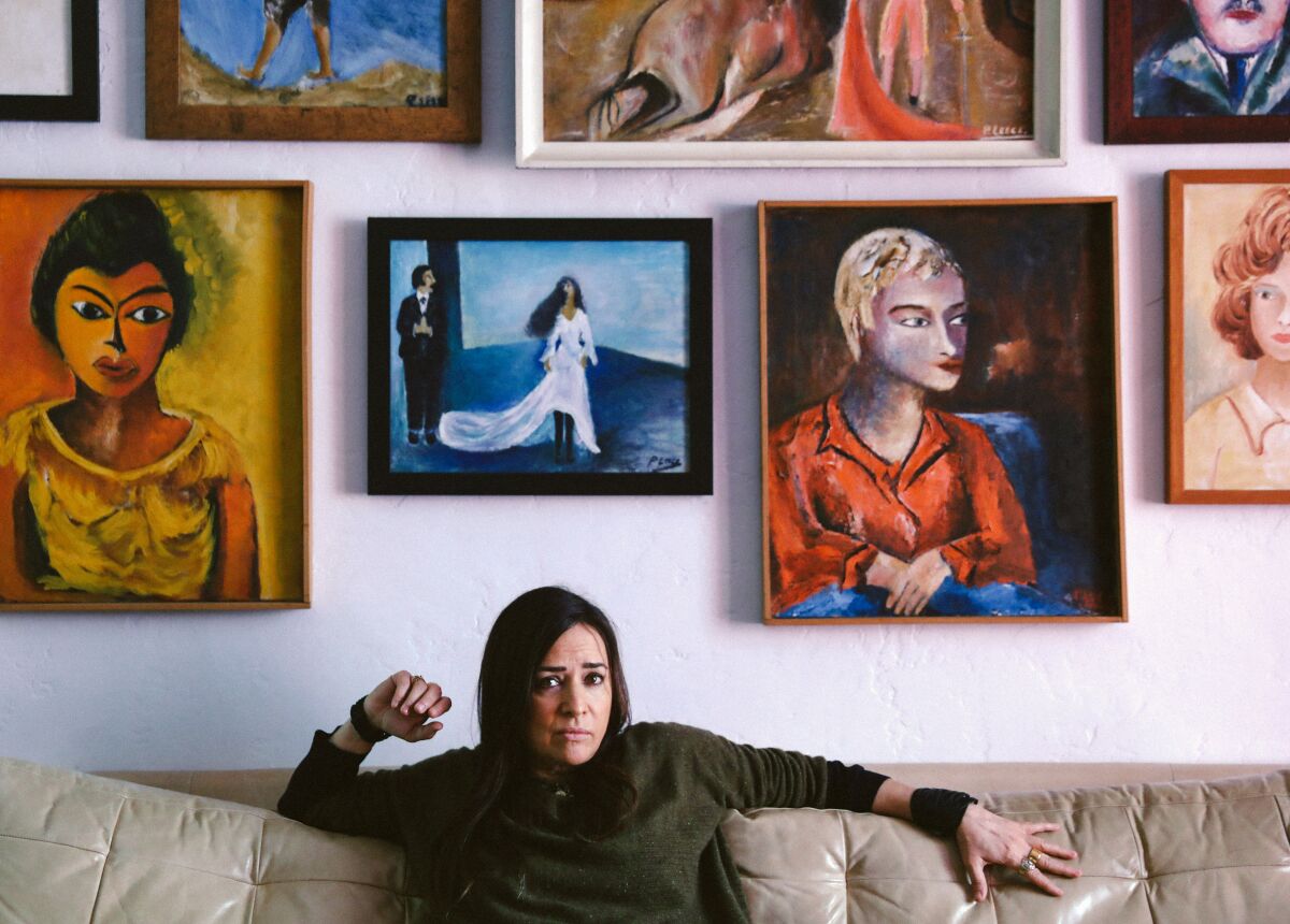 A woman sitting on a couch with a wall of paintings of women's faces above her.
