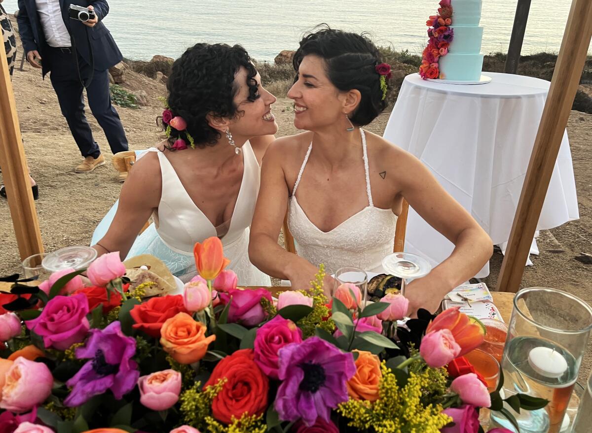 Two women in white leaning in to one another as they sit at a table on a beach, with colorful flowers in the foreground
