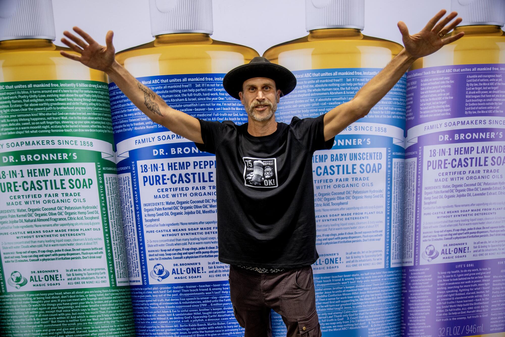 A man in a hat standing and raising both arms in front of an enlarged photo of bottles of Dr. Bronner's soap.