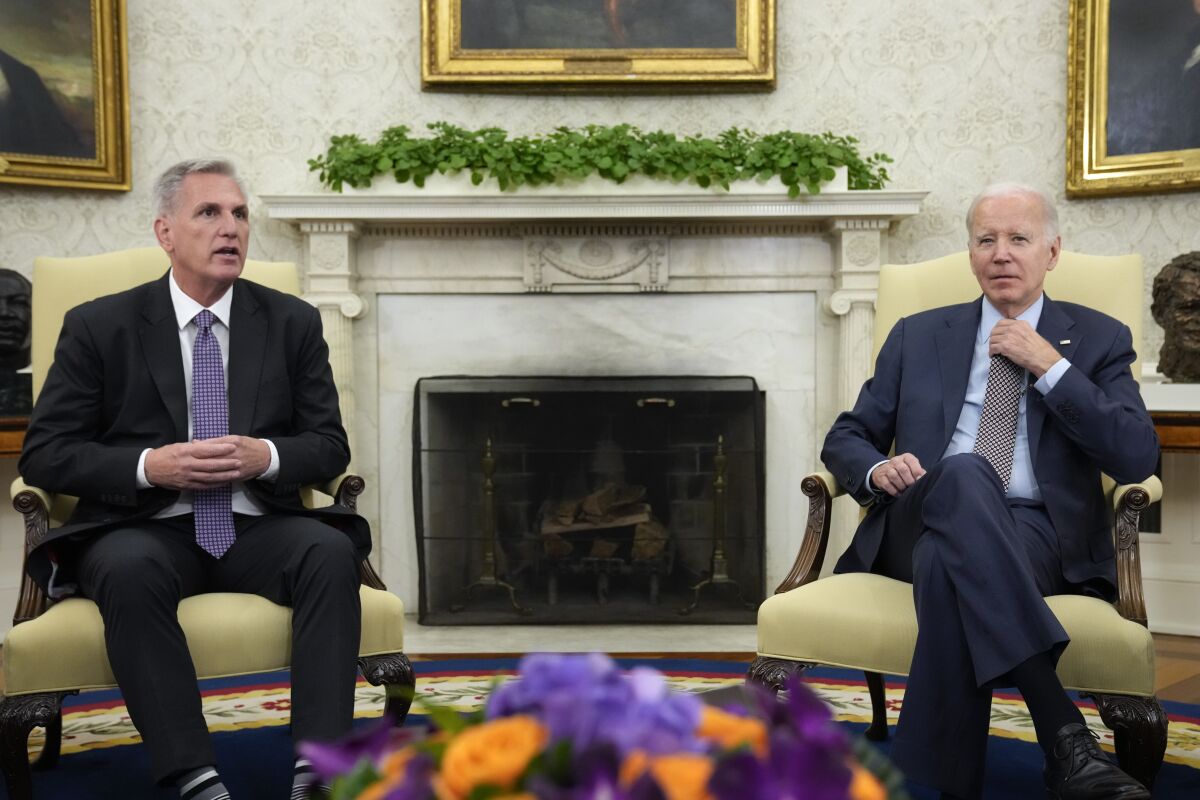 House Speaker Kevin McCarthy and President Biden, both wearing dark suits, are seated in armchairs flanking a fireplace.