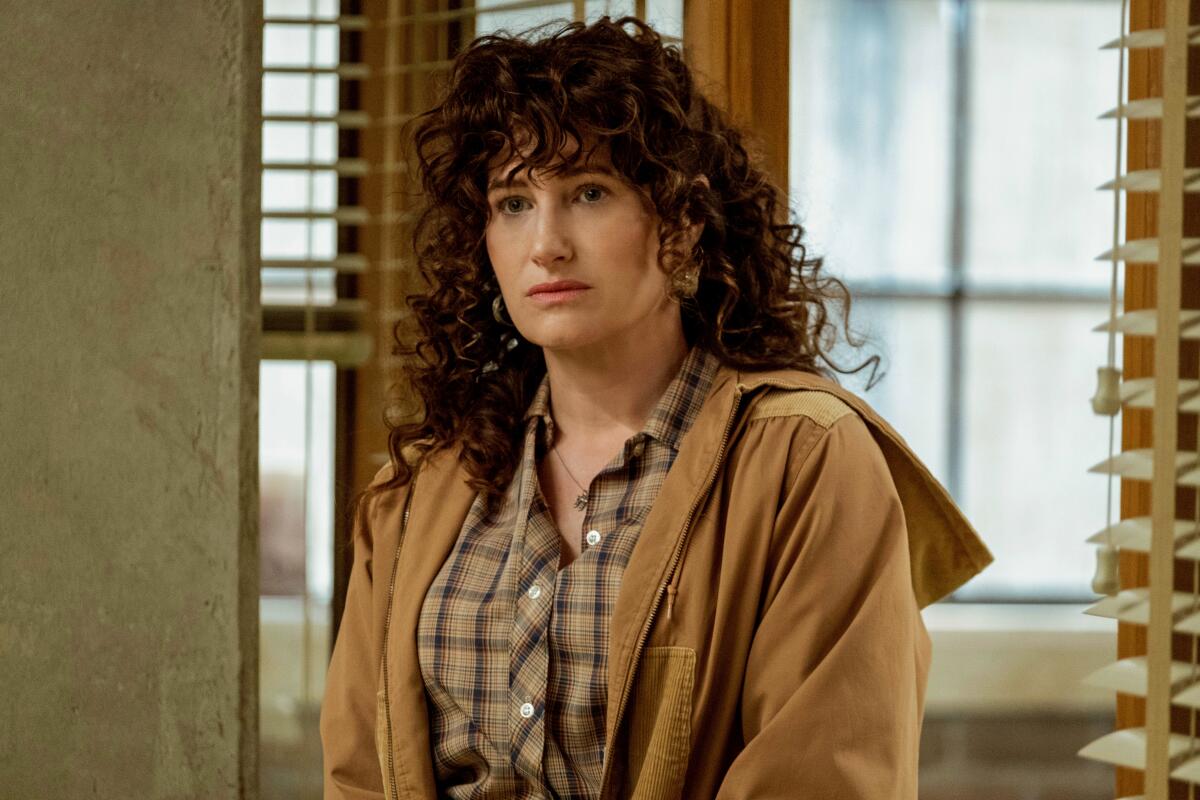 A woman with curly brown hair in a brown jacket and plaid shirt