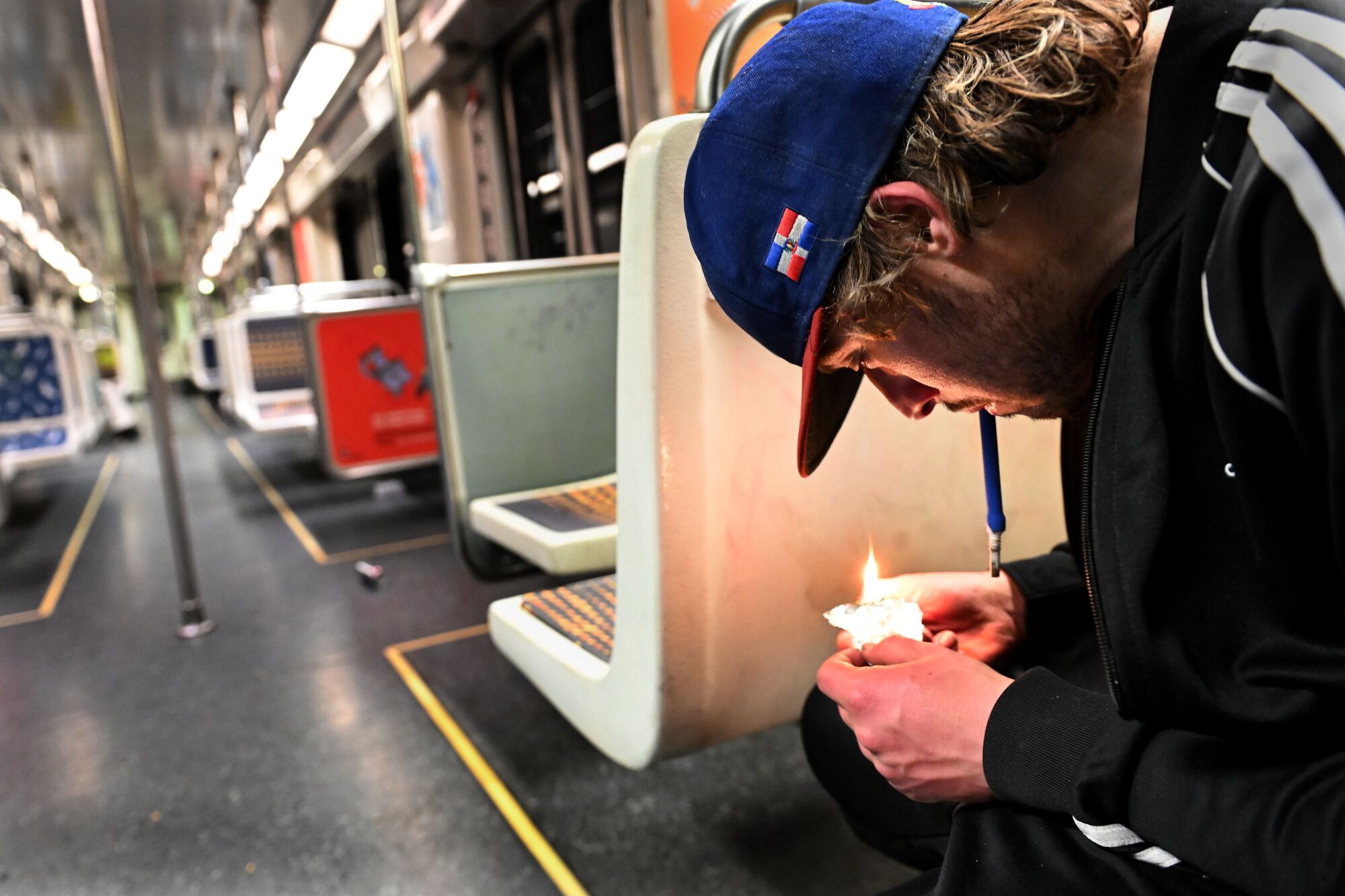 Seated in a subway train, a young man with dark blond hair and a baseball cap smokes a substance off of an aluminum foil.
