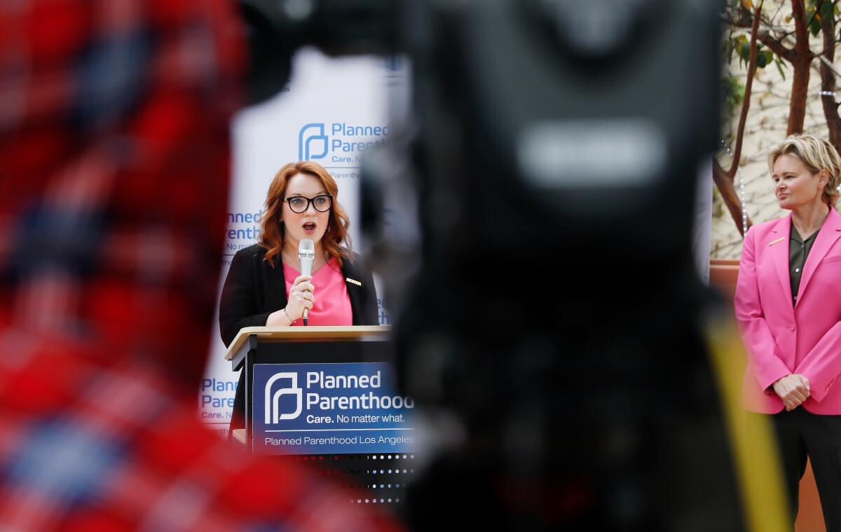 Lindsey Horvath, wearing a pink shirt, speaks at a lectern with a Planned Parenthood sign; nearby, a woman in a pink blazer