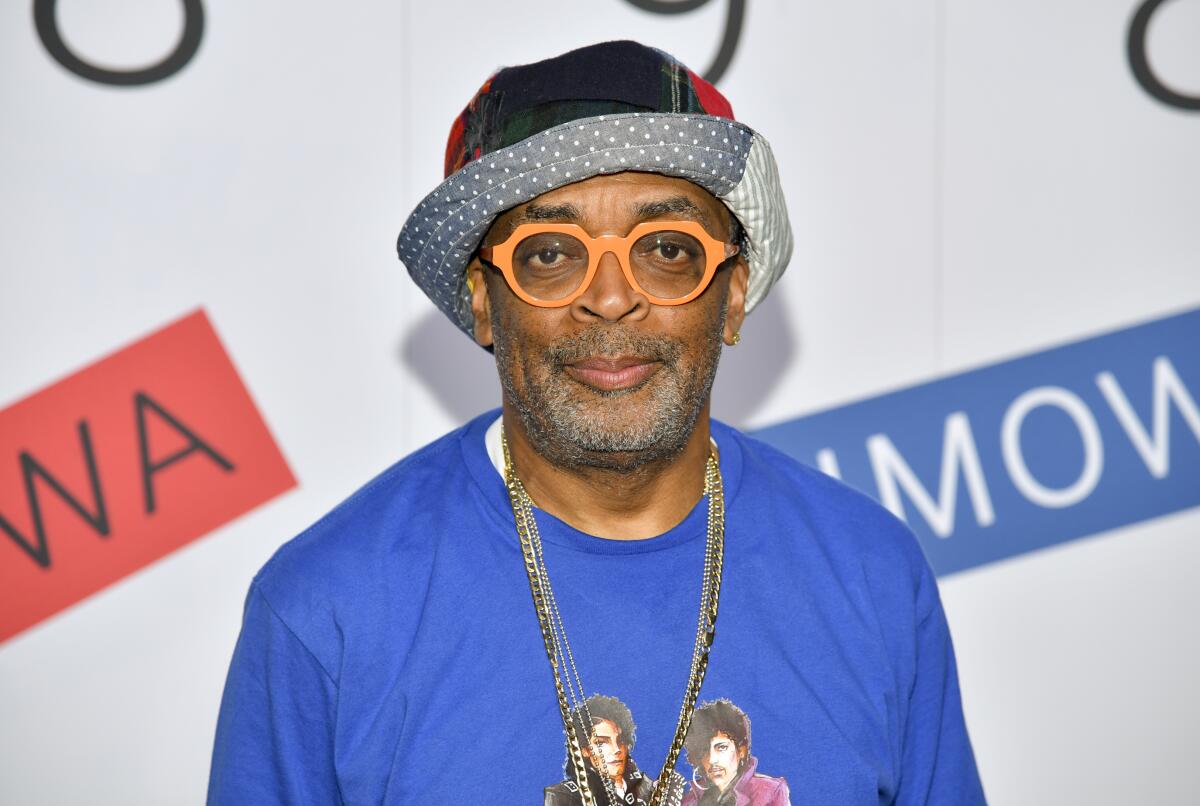 A man in orange glasses, a patterned hat and a blue shirt with photos of Michael Jackson and Prince, looks straight ahead.