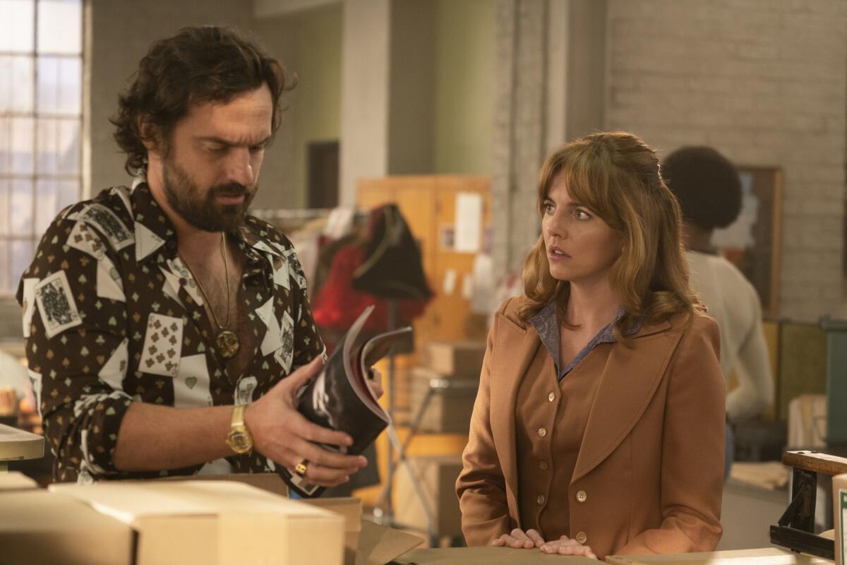 A man in a patterned shirt holding a magazine. To his right, a woman in a brown shirt and jacket looks at him