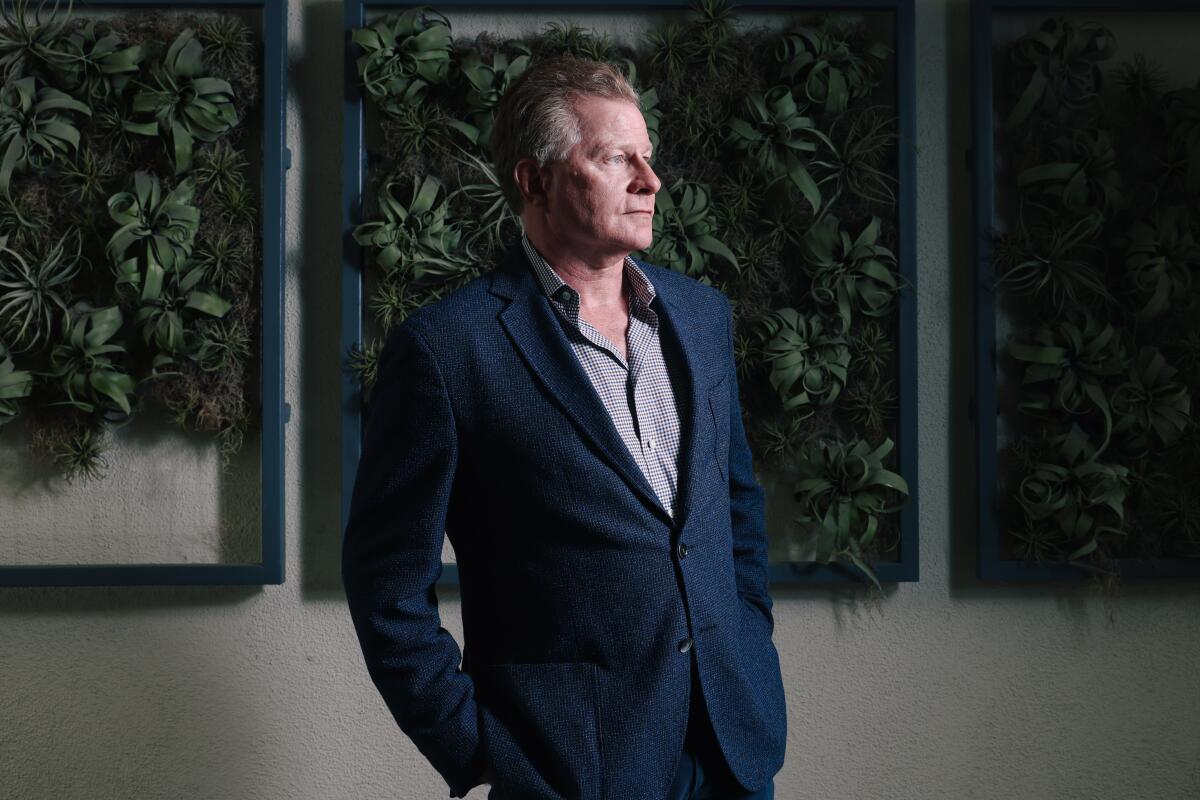 Screenwriter David Magee wears a blue suit and stands near a wall of foliage for a portrait.