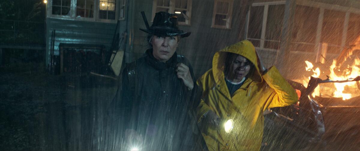 A woman with a rifle and a woman with a flashlight in the rain beside a burning vehicle in the movie "Lou." 