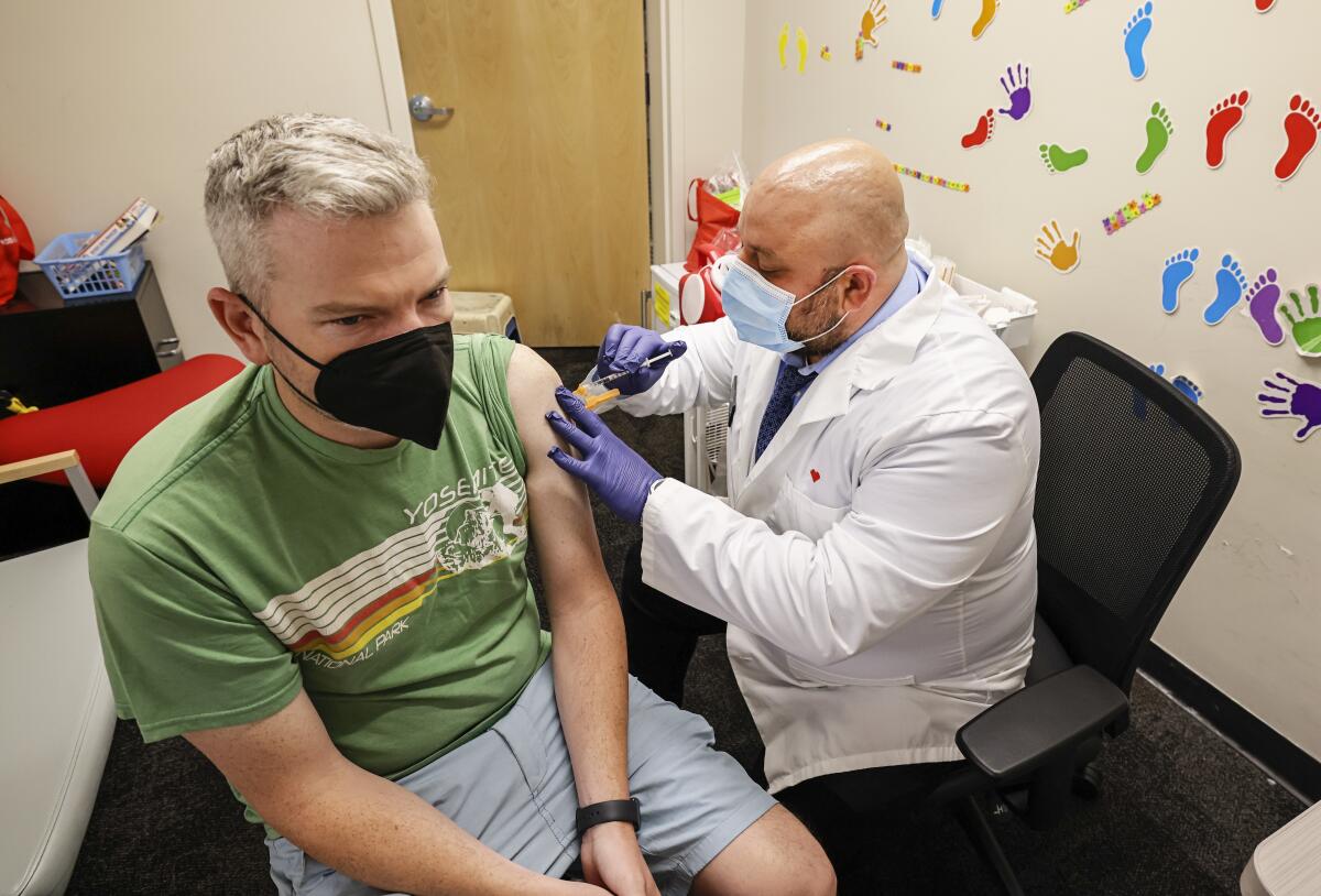 A man wearing a white coat, gloves and a surgical mask gives a man wearing a Yosemite T-shirt and a mask a shot in his arm.