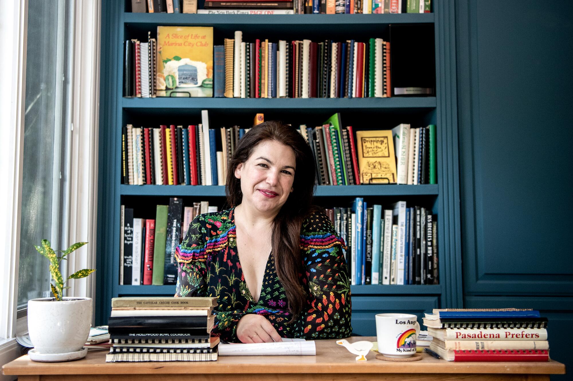 A woman sits at a desk in front of a large shelf of cookbooks.