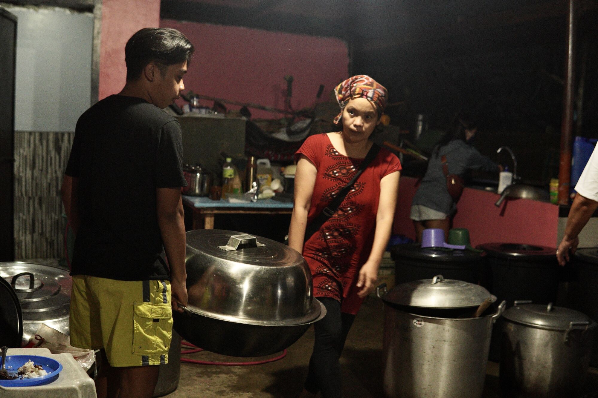 Two people carry a big metal pot in a room with other big pots.