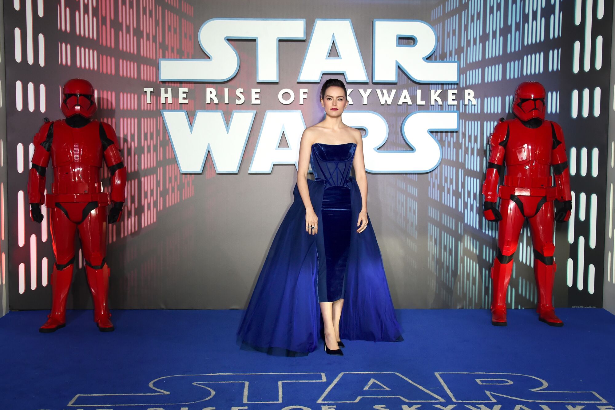 Une femme brune vêtue d'une robe corsage bleue devant un panneau Star Wars.