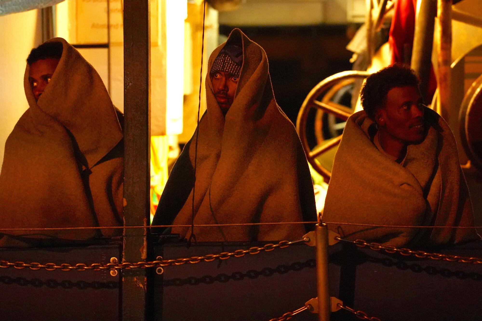 Three men wrapped in blankets on a boat.