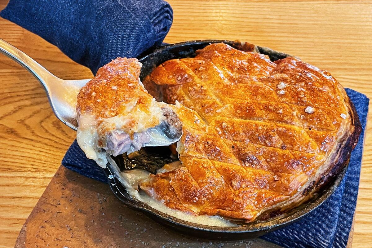 Duck pot pie in a skillet, its crust a puffy browned pastry dough. A silver spoon lifts a bite from the side.