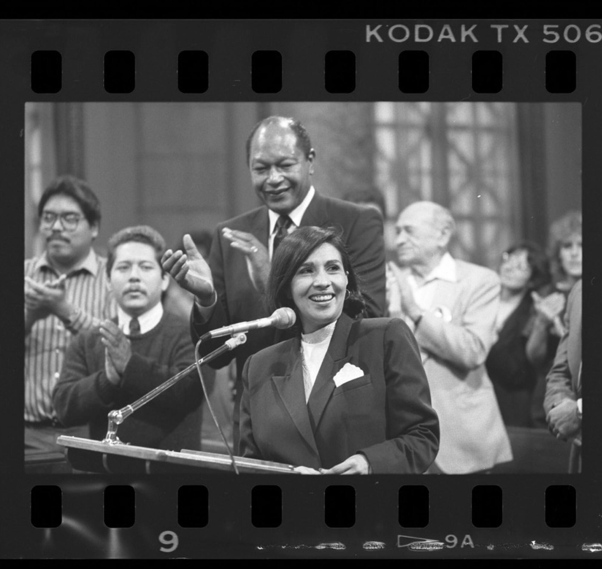 In a black and white photo frame, a woman stands at a lectern, with people behind her clapping.