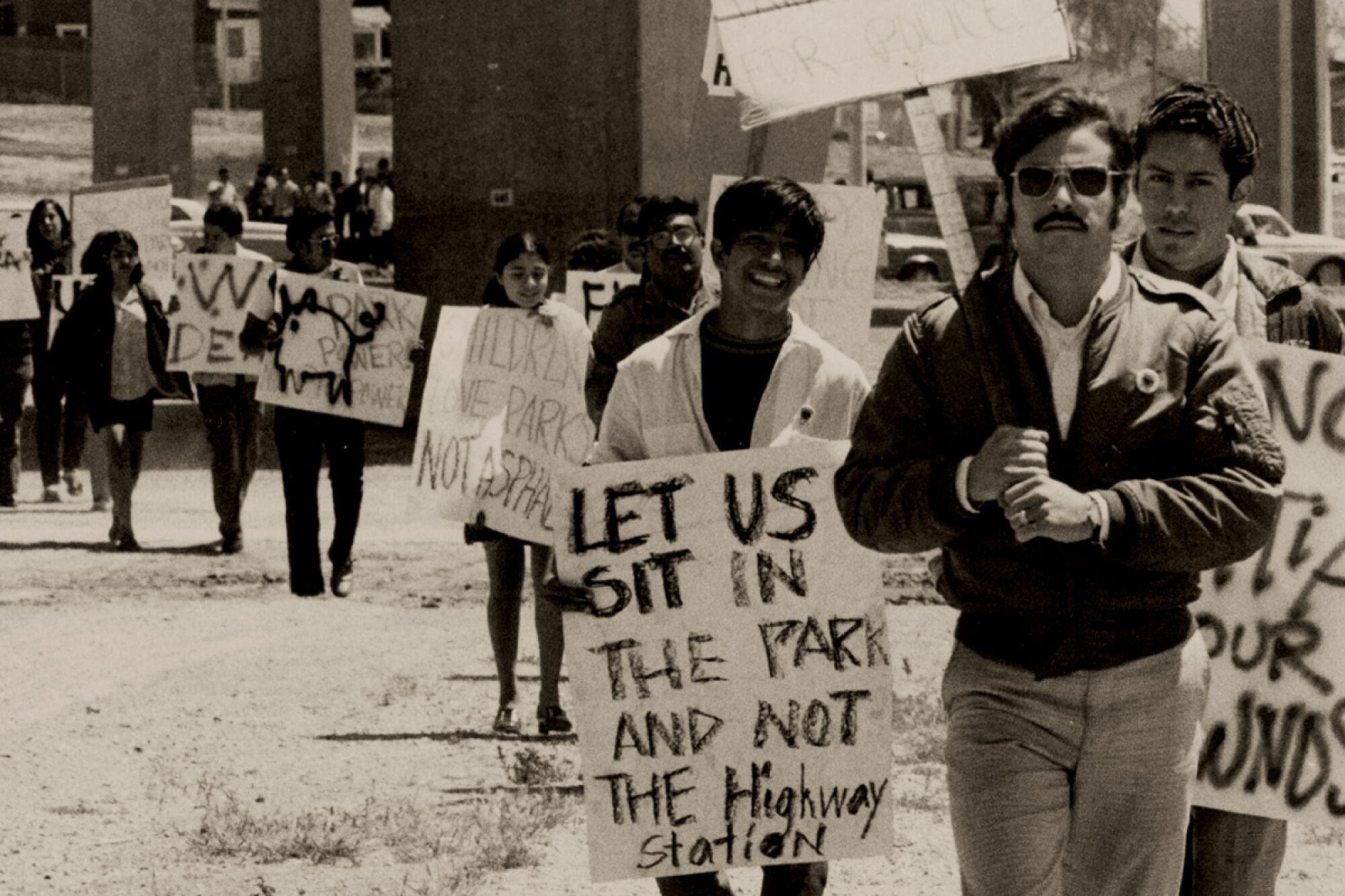 Black-and-white photo of protesters carrying signs with messages like "Let us sit in the park and not the highway station."