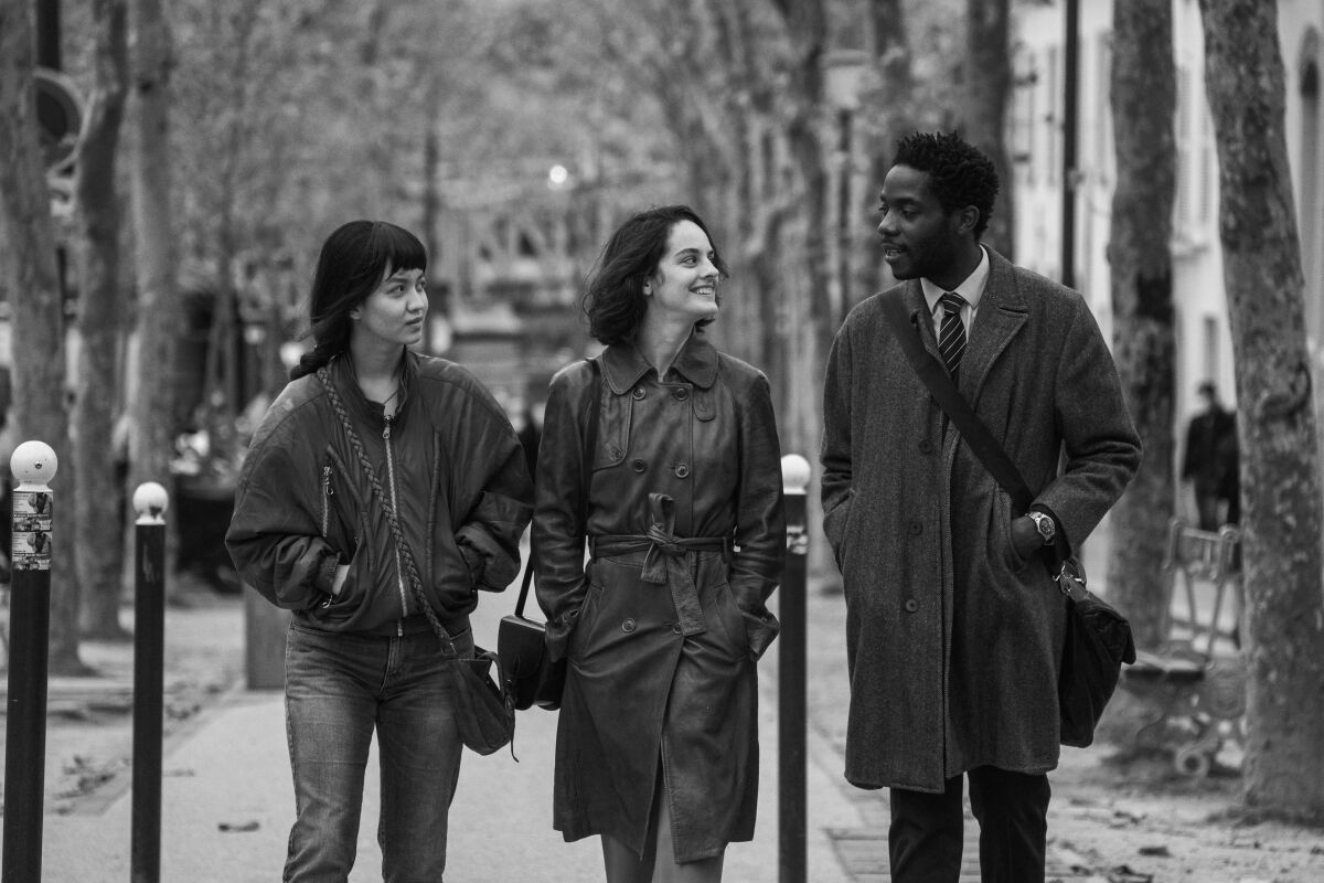 Two women in light coats and a man in a tie and trench coat talk and walk down a city street bordered by trees.