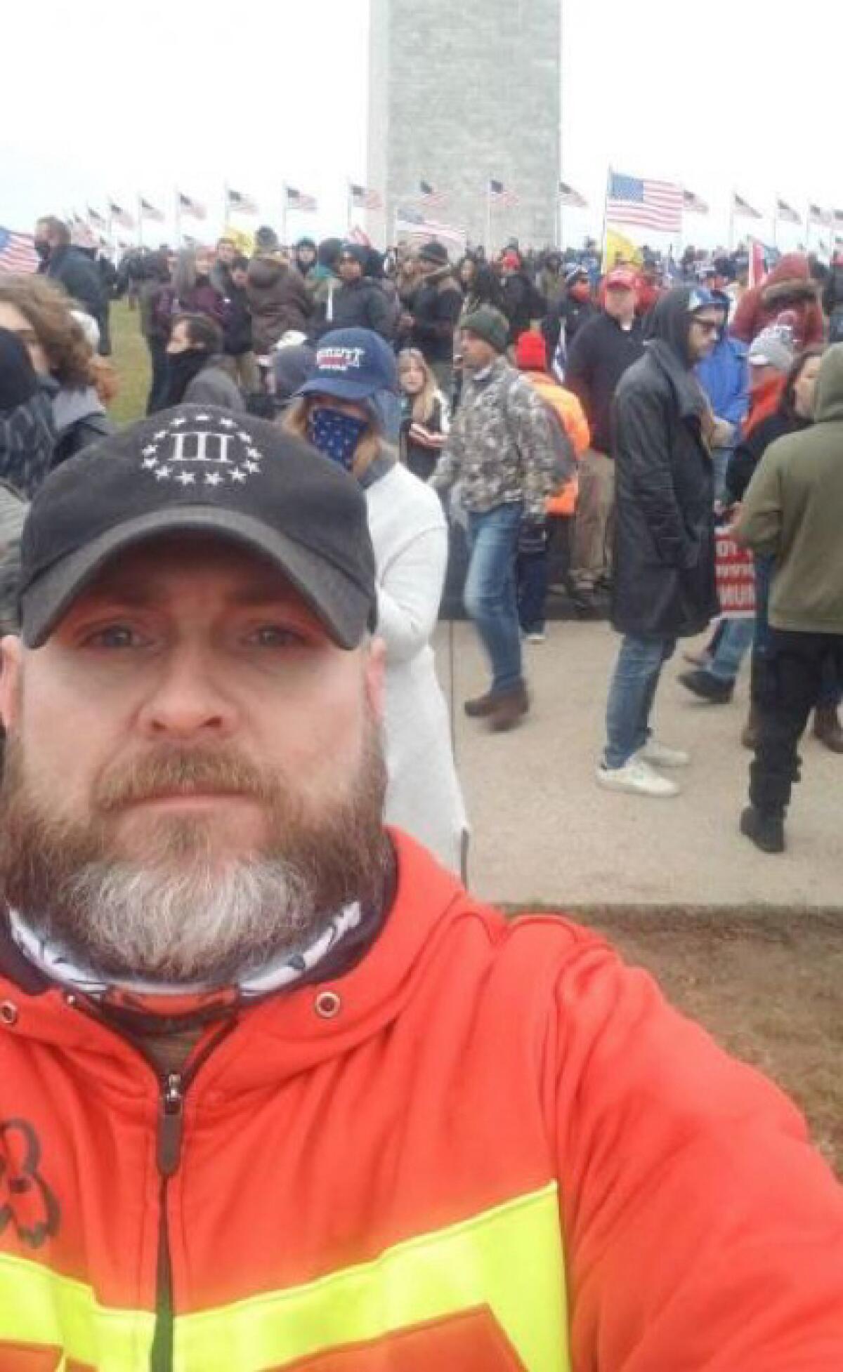 A bearded man in a dark baseball cap pictured from the chest up, with a crowd and a line of American flags in the background.