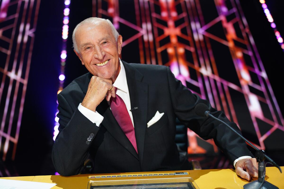 An older man in a suit sits at a table and smiles while resting his chin on his right hand