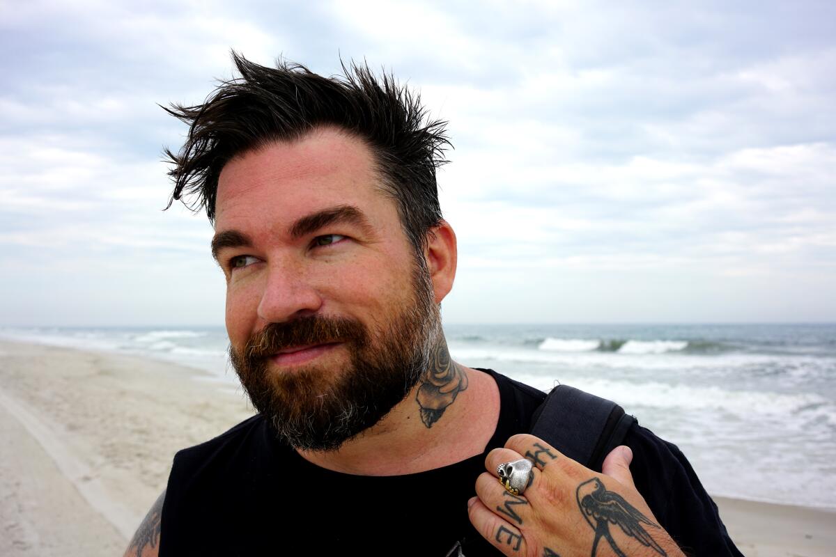 A tattooed man in a black shirt stands on the beach with the ocean behind him.