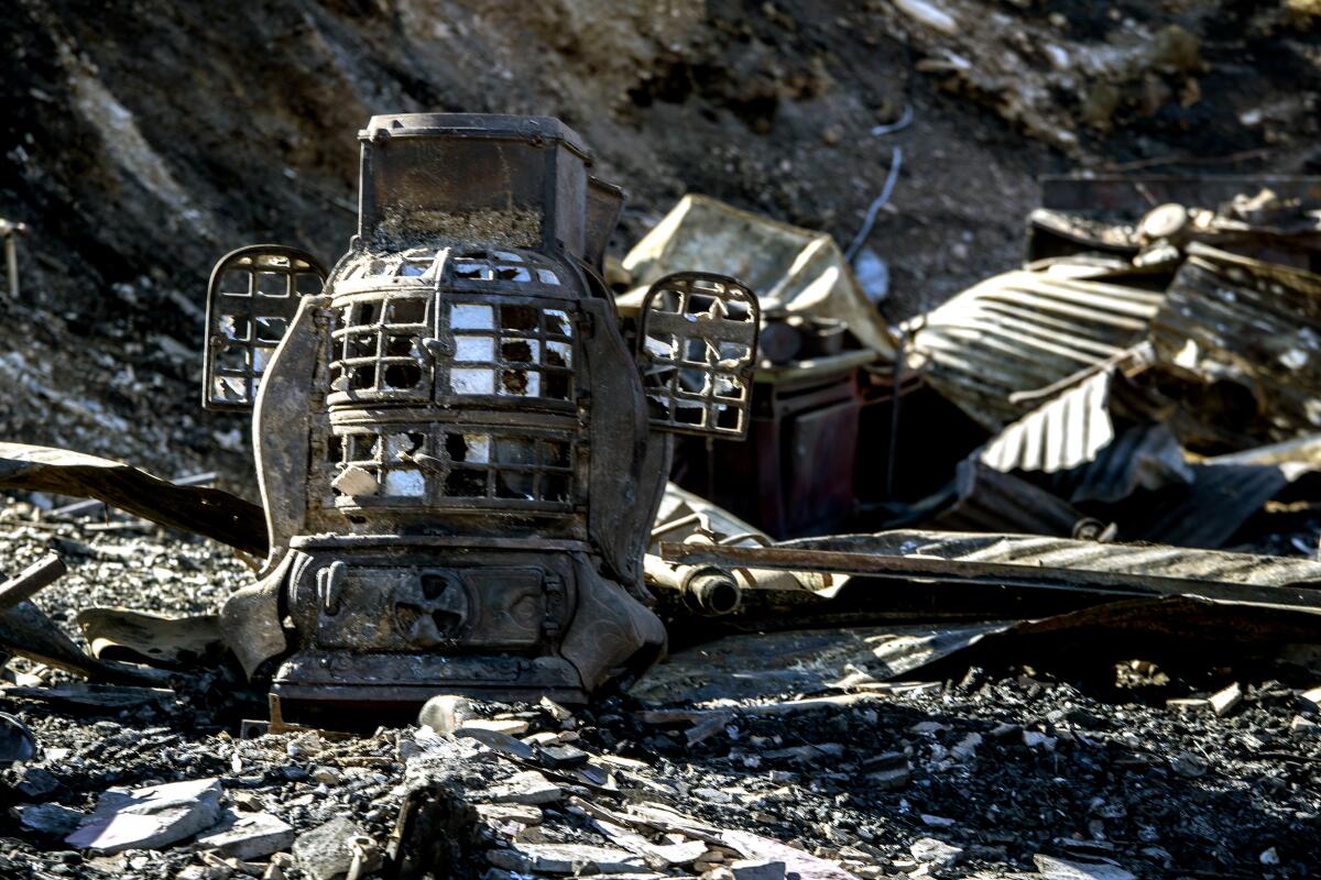 A fire-blackened old stove with broken glass panes and two small doors ajar sits among other charred debris