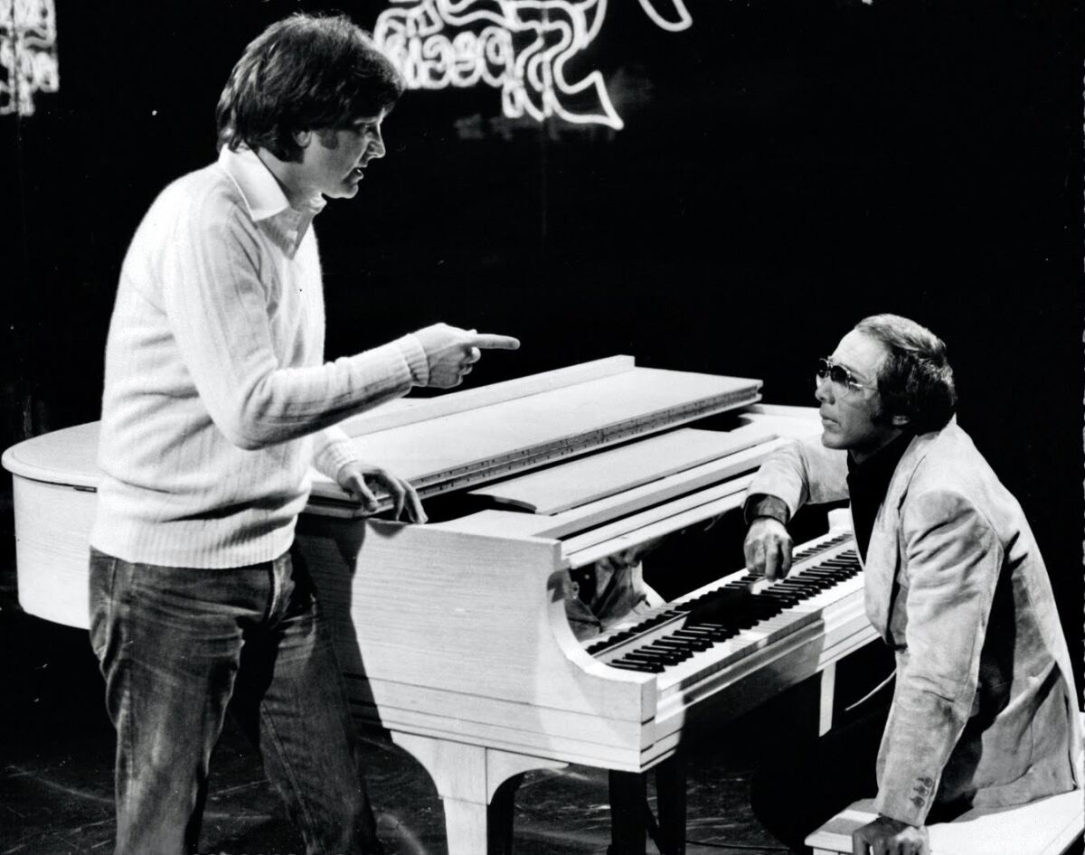 Two men by a white grand piano.