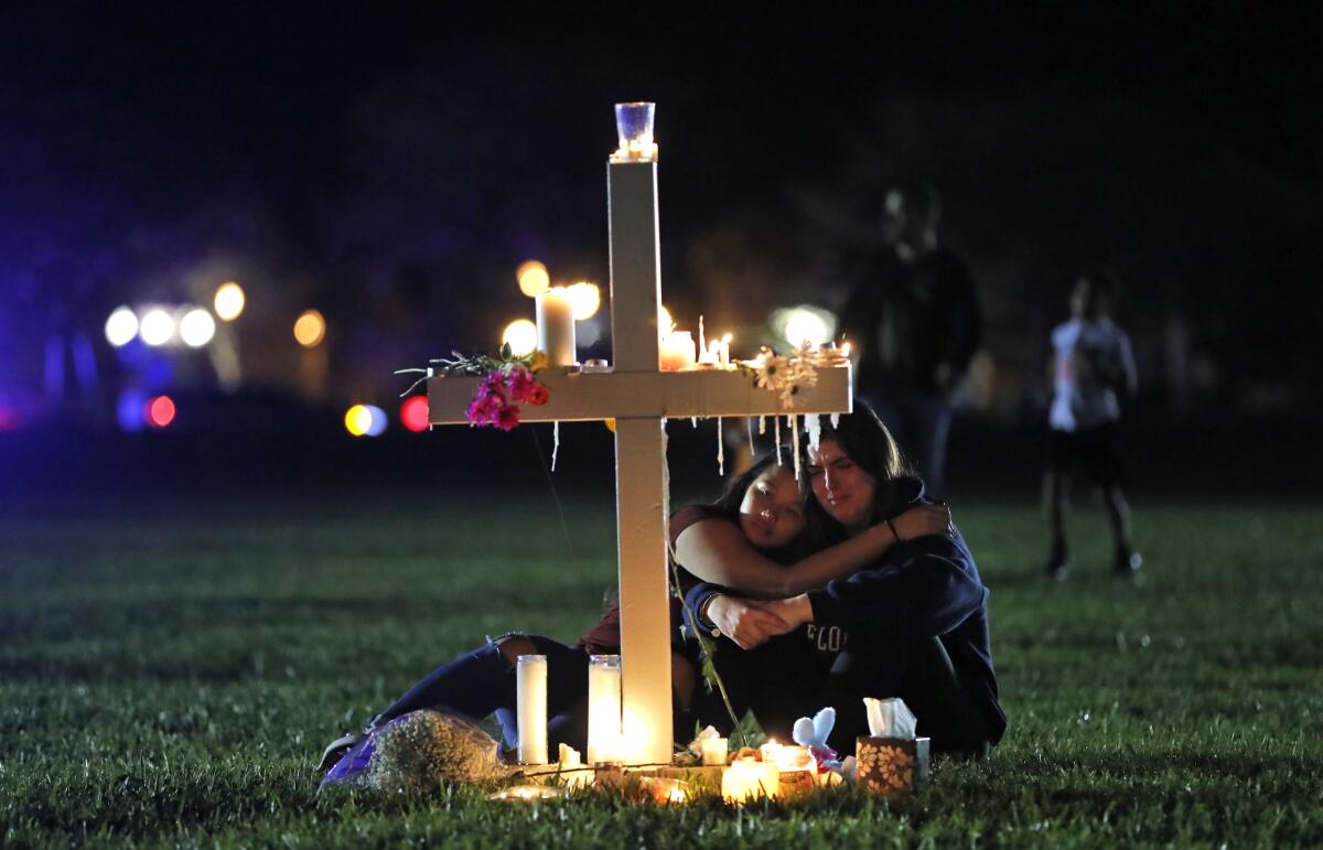 People lean on each other as they sit by a memorial cross lit by candles. 