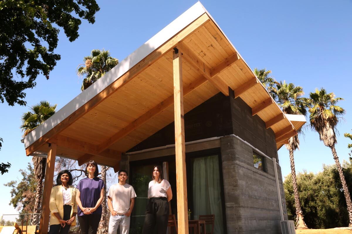 Four people stand on the front porch of a small concrete house with a dramatic sloping roof made of wood.